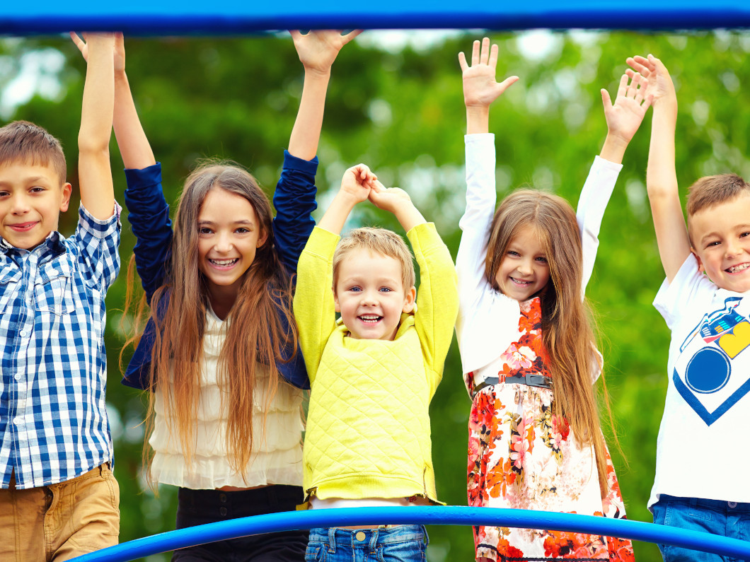 imagen en la que aparecen 5 niños y niñas sonrientes mirando a cámara, con los brazos levantados. Están jugando en un parque infantil