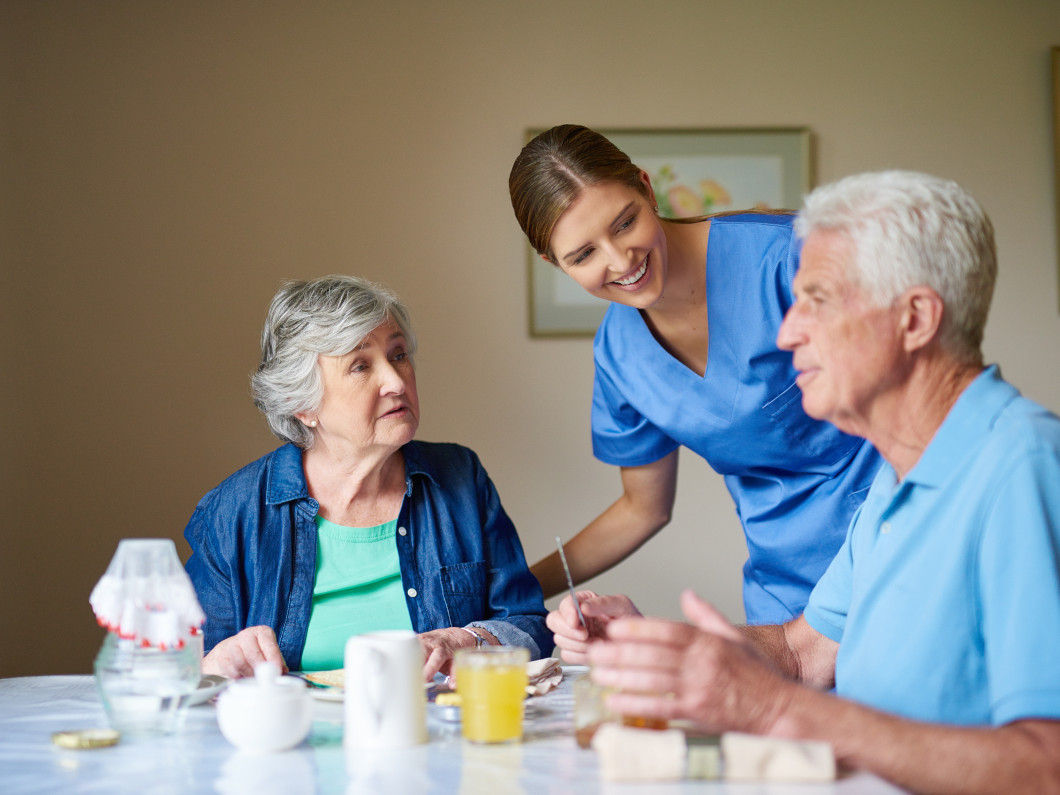 imagen de una pareja de personas senior que desayunan en una residencia de mayores, acomopañados por una sanitaria que les ofrece su ayuda