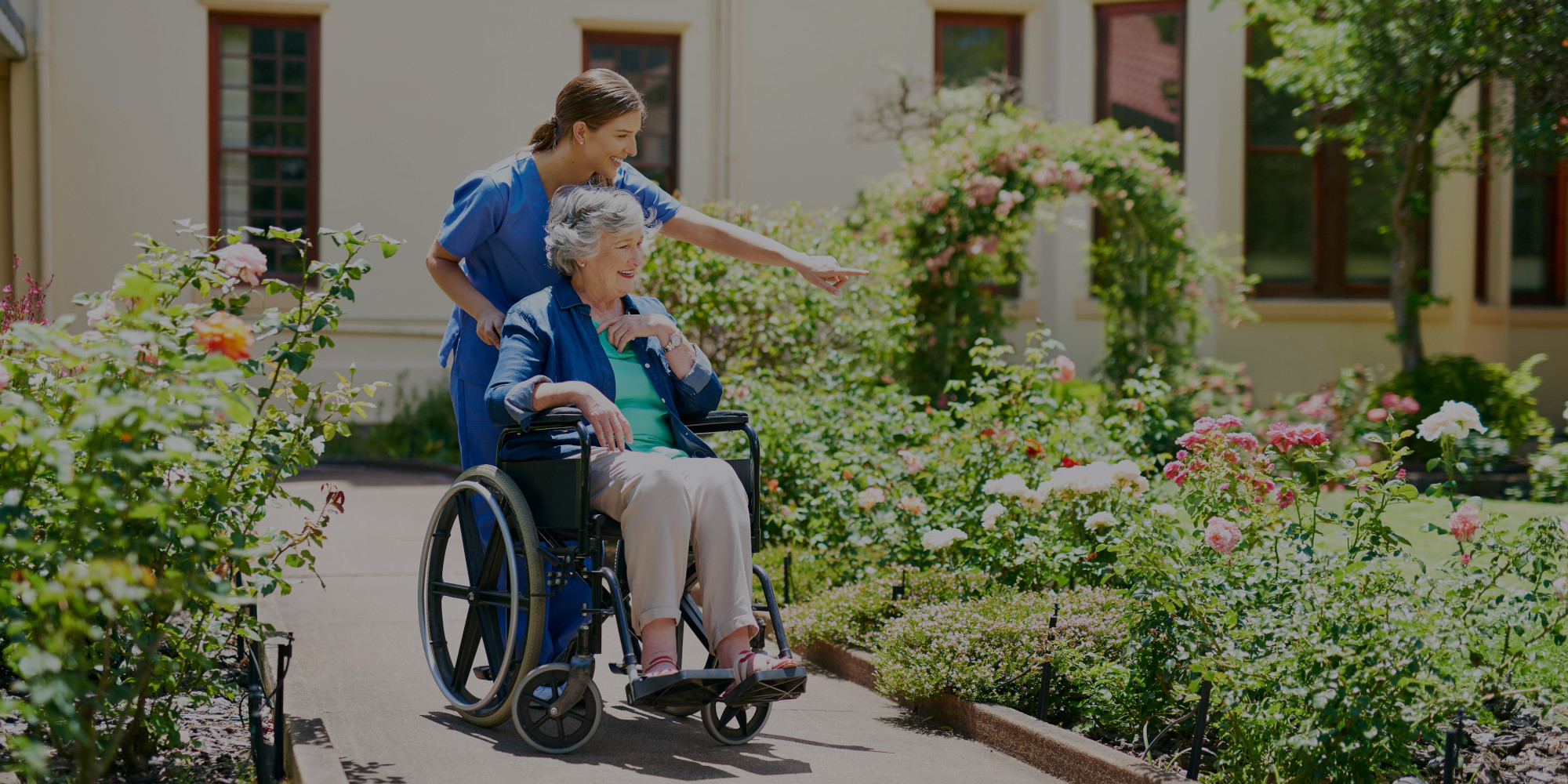 imagen de una mujer de avanzada edad que está sentada en una silla de ruedas paseando junto a una mujer sanitaria por unos jardines un día soleado