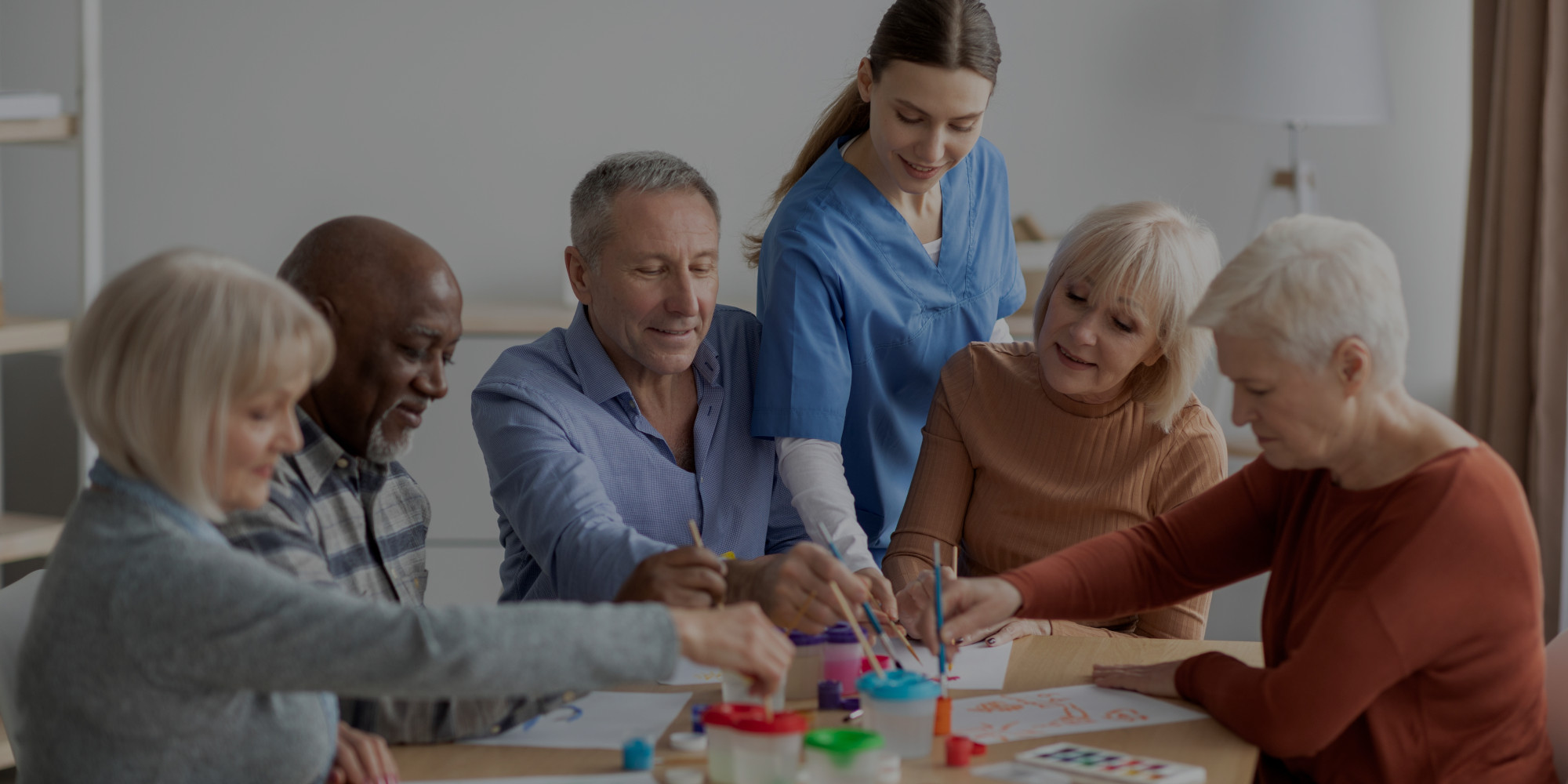 imagen de un grupo de personas adultas, mayores de 60 años, que están realizando manualidades alrededor de una mesa. Están acompañados por una mujer sanitaria